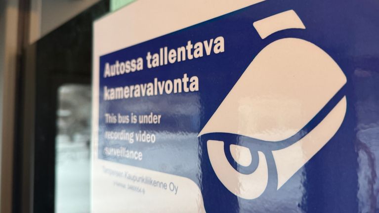 Kuvassa näkyy kaksikielinen ilmoitus, joka kertoo, että bussissa on tallentava kameravalvonta. Teksti on suomeksi ja englanniksi.