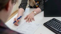 En person fyller i ett formulär medan en annan person pekar på papperet vid ett skrivbord med en dator och ett tangentbord.