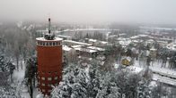 Vanha, punatiilinen vesitorni keskellä lumista mäntymetsää, taustala näkyy Heinolan kaupunki. 