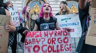 En kvinna med rosa hår håller i en skylt under en protest där det står "ta hand om min våldtäktsmans bebis eller gå till skolan?"