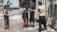 A group of five kids spraying water with a hose on a rather run-down sidewalk in bright sunlight.
