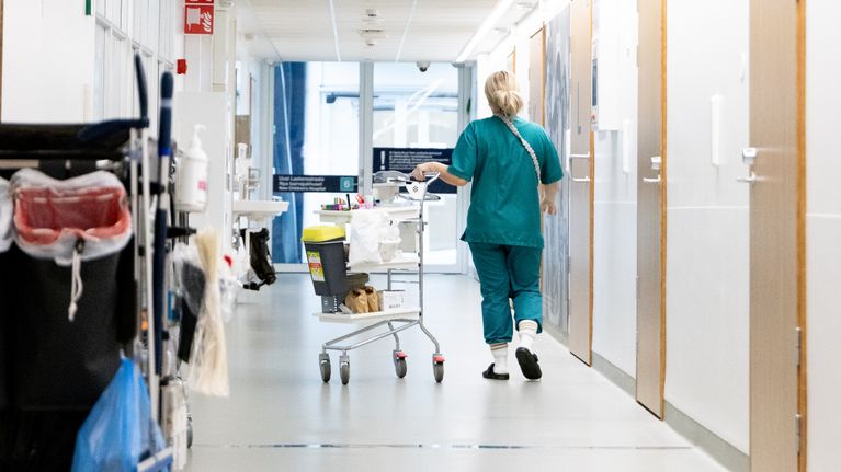 Sairaalan käytävällä työntekijä vihreään sairaalan asuun puketuneena työntää kärryä.