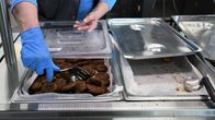 Kaksi isoa tarjoiluastiaa suurtalouskeittiössä, toinen sisältää falafelpyöryköitä ja toinen on tyhjä. Siniseen kumihanskaan puettu käsi järjestelee metallilastalla pyöryköitä.