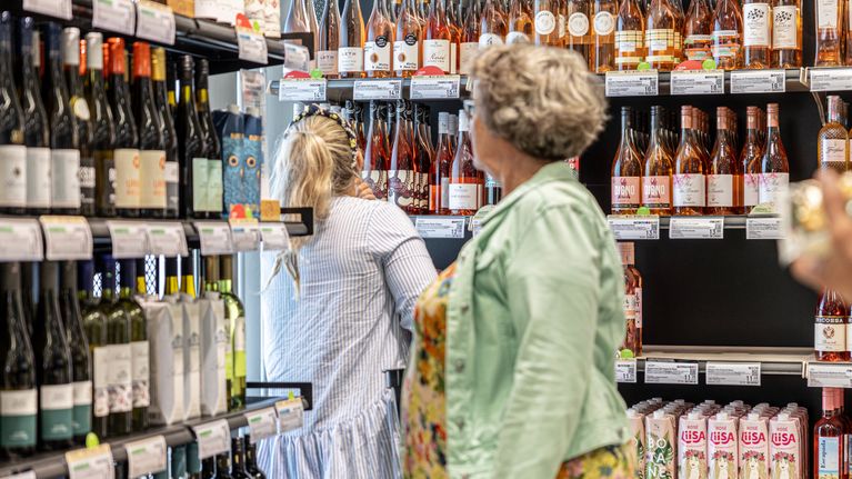 Two women in a wine store aisle, with shelves filled with wine bottles.