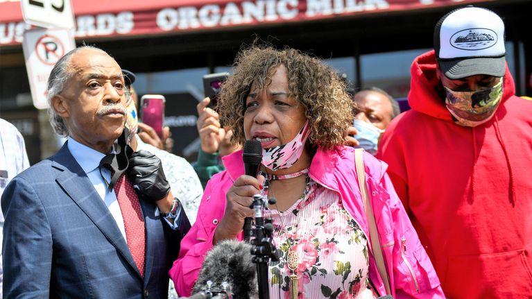 Eric Garnerin äiti Gwen Carr puhuu mikrofoniin. Al Sharpton kuuntelee vakavana vieressä. Taustalla näkyy väkijoukkoa ja elintarvikekaupan julkisivua.