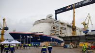 A blue and white ship with the name Mardi Gras in orange, people walking and on bikes in blue and yellow construction outfits in the foreground on a grey day.