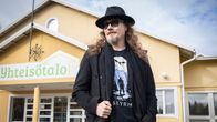 Mustaan lierihattuun ja takkiin pukeutunut pitkätukkainen mies seisoo keltaisen rakennuksen edessä.