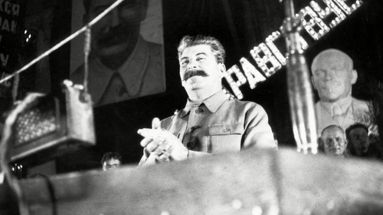 Stalin seisoo korokkeella hieroen käsiään yhteen viiksekkäänä ja univormussa. Taustalla vasemmalla näkyy suuri Stalinin ja oikealla Stalinin selän takana Leniniä esittävä patsas. 