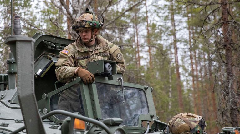 Sotilas seisoo panssariajoneuvon kyydissä metsässä. Hänellä on Yhdysvaltojen lippu olkavarressa.
