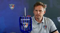 Päävalmentaja Vesa Vasara harmaassa FC Inter Turku -paidassa istuu sinisen taustan edessä, pöydällä pieni FC Inter Turku -viiri.