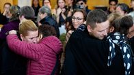 Useat ihmiset halaavat toisiaan sisätiloissa. Taustalla on muita henkilöitä seuraamassa tilannetta. 