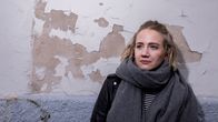 Anniina nahkatakissa ja paksussa harmaassa kaulahuivissa, kuvattu vasten seinää, josta lohkeillut valkoista maalia.