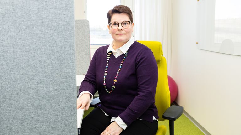 Henkilö katsoo kameraan ja hymyilee tietokonetuolilla istuessaan toimistotilassa.