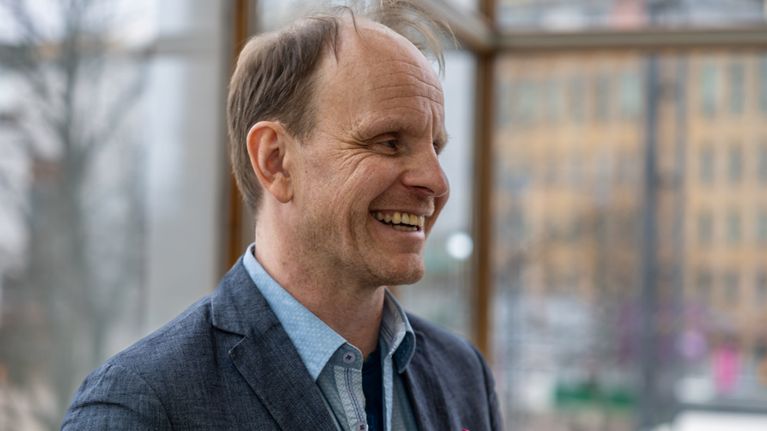 Dome Karukoski hymyilee rennosti ja katsoo poispäin. Pikkutakin taskussa lila liina.