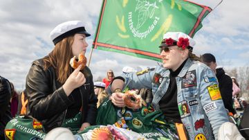 Kaksi opiskelijaa syö munkkeja istualtaan ylioppilaslakit päässä, taustalla liehuu metsäylioppilaiden lippu.