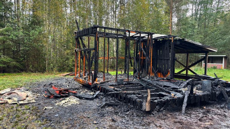 Ett utbränt trähus står kvar i en glänta omgiven av träd, med svarta, förkolnade stolpar. Väggarna är borta.