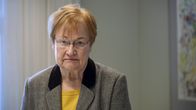 An elderly woman in a suit jacket, looks seriously into the camera.