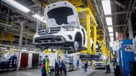 Autotehtaalla työntekijät kokoavat autoja tuotantolinjalla, jossa useita autoja roikkuu nostureilla samanaikaisesti.