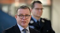 A man with brown hair, round brown glasses and a dark suit speaks with a blurry man in uniform behind him.