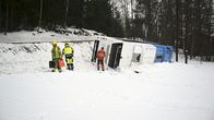 Linja-auto on kaatunut ojaan lumisessa maisemassa tien vieressä. Huomiopukuiset ihmisiä auton ympärillä.