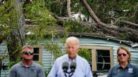 En suddig Joe Biden i förgrunden, i bakgrunden träd som fallit över ett hus.
