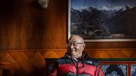 Kanchha Sherpa istuu toppatakki yllään. Hänen takanaan seinällä on taulu vuorista.