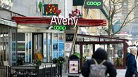 Kaksi henkilöä kävelee kadulla Coop-kyltin ja Aveny-ravintolan edustalla, taustalla terassikalusteita.