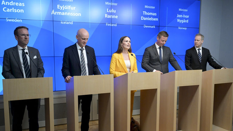 Four men in dark suits and a woman in a yellow blazer between them, standing in a row at podiums, at a press conference.