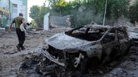 A man holding a dog walks through the aftermath of a bombing, with a destroyed car in the foreground and trees and a building in the background.