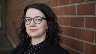 Saara Loukola seisoo punatiilistä seinää vasten pukeutuneena mustaan pukuun ja kuvioituun puseroon.