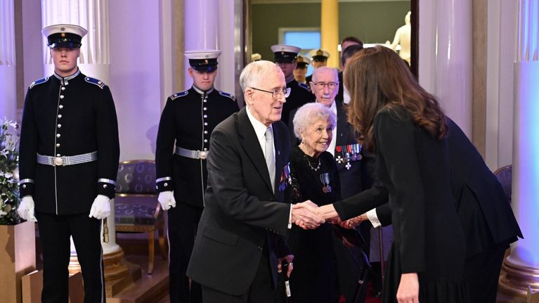 Presidentparet välkomnar veteraner och lottor till slottet: en mörkkläd Suzanne Stubb böjer sig framåt för att skaka hand med en man i mörk kostym med ett stort antal ordnar på bröstet.
