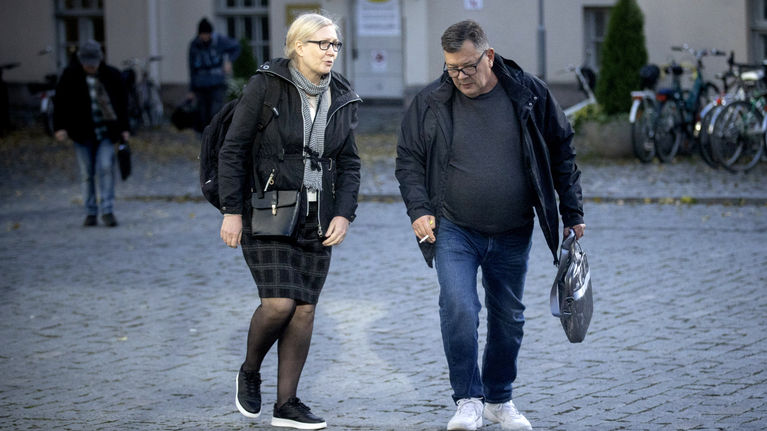 Middle aged woman and man carrying bags walking on a brick road on a grey day, with a building and a few people in the background.
