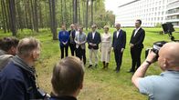 Ryhmä ihmisiä seisoo rivissä nurmikolla metsän ja suuren valkoisen rakennuksen edessä, useita valokuvaajia kuvaa heitä.