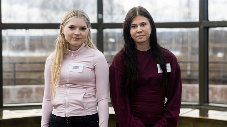 Kaksi nuorta seisoo vierekkäin sisätiloissa suurten ikkunoiden edessä nimilaput rintapielessään.