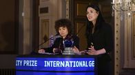 Two women with dark hair and dark clothing at a podium, one is speaking into a microphone.