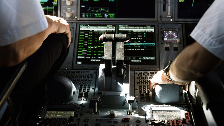 Arms of two pilots seen from behind in a dark airplane cockpit with many buttons and screens.