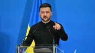 A man with dark hair and beard, wearing a black shirt and pants, standing at a podium and pointing with his left finger. The background is blue.