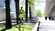 Henkilö haravoi kävelykatujen välistä viheraluetta huoltotyövaatteissa.