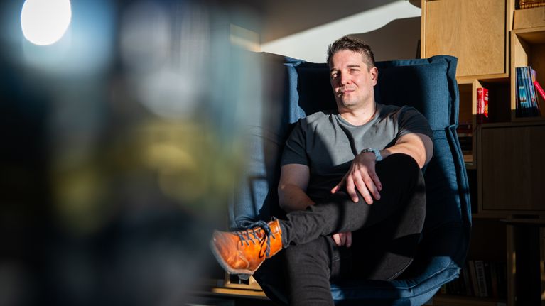 Mies istuu mukavasti sinisellä nojatuolilla, jalat ristissä, pukeutuneena rentoon harmaaseen t-paitaan ja mustiin housuihin. Valo luo huoneeseen lämpimän ja kodikkaan tunnelman.