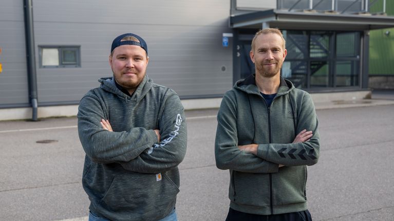 Två män med armarna i kors står bredvid varandra framför en idrottshall.
