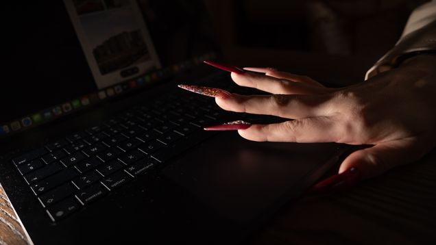 Punaisten akryylikynsien koristama käsi koskettaa kannettavan tietokoneen kosketuslevyä. Aurinko paistaa kuvakulmaan, luoden tyylikkäitä varjoja käsivarren ja näppäimistön ylle.