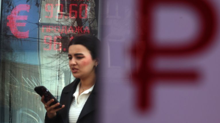 En person håller i en mobil framför en valutakursdisplay med texten om valutakurs och en röd euro-symbol.