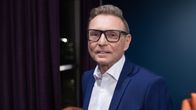 Mies pukeutuneena tyylikkääseen siniseen pukuun ja valkoiseen kauluspaitaan seisoo huoneessa, jonka taustalla näkyy tummia verhoja ja oranssi tuoli.