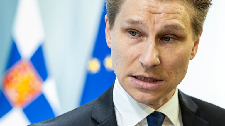 Headshot of a man in a dark suit and necktie, with Finnish and EU flags hanging in the background.