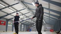 Kaksi talviliikuntatamineisiin sonnustautunutta nuorta miestä seisoo curlingharjat käsissään hallissa jäällä, johon on maalattu curling- kentät.