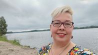 Kati Tialan omakuva, jossa hän hymyilee. Tialalla on vaaleat lyhyet hiukset, silmälasit ja värikäs paita. Taustalla järvi.