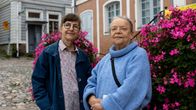 Två personer står på en kullerstensgata mellan två stora blomkrukor med rosa blommor, hus och museiskylt i bakgrunden.