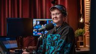 En person med hörlurar och mikrofon står vid ett radiobord, datorskärmar och studioteknik syns i bakgrunden.