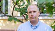 Sinipaitainen mies seisoo keltaisen talon edustalla vihreiden puiden katveessa.