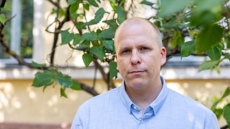 Sinipaitainen mies seisoo keltaisen talon edustalla vihreiden puiden katveessa.